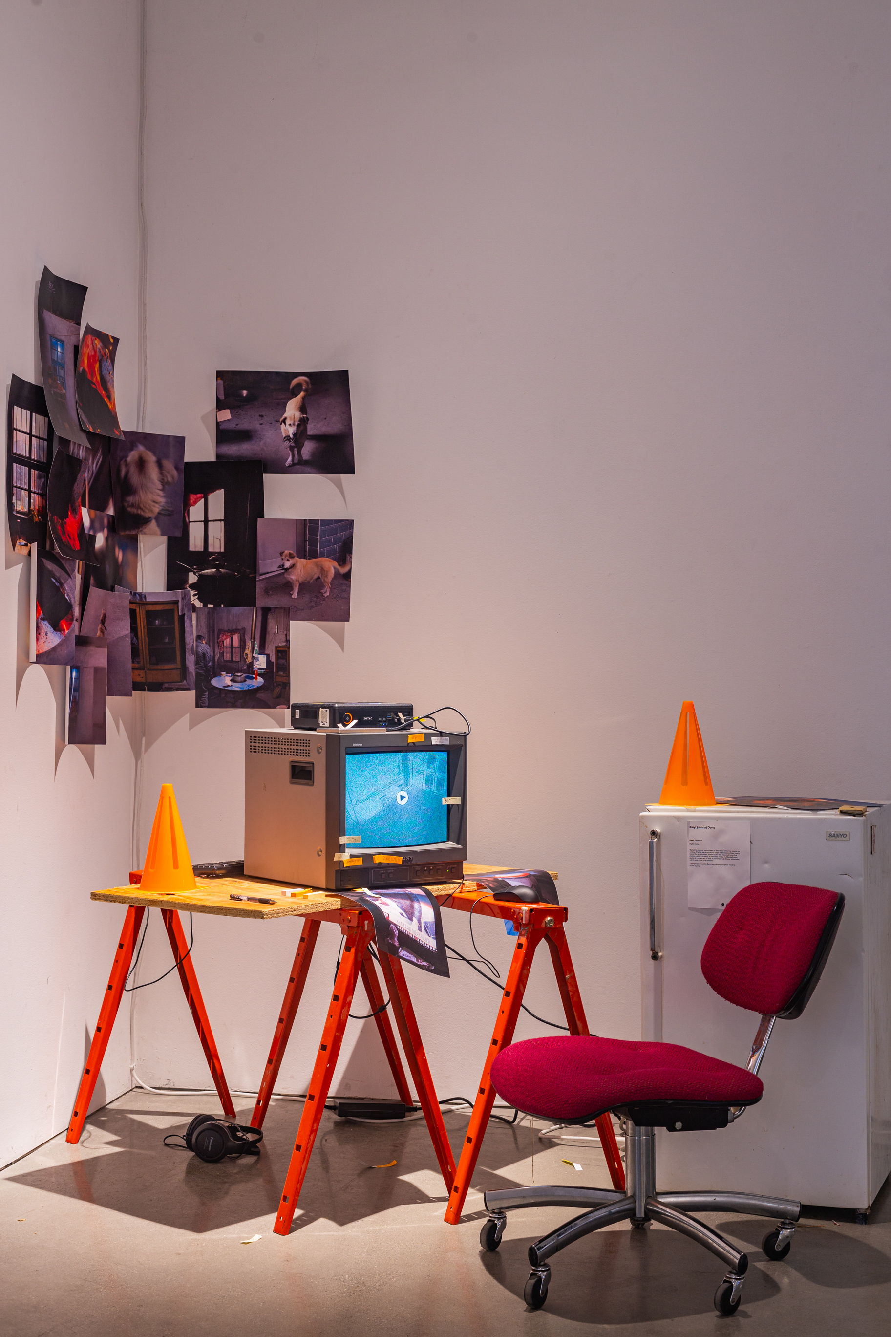 A sculptural piece, on the right is a white fridge with a plastic cone on top of it, in the center is a makeshift construction desk with plywood and an office chair, on top of it is a CRT tv with visuals on it, in the background is printed posters of photogrpahy.